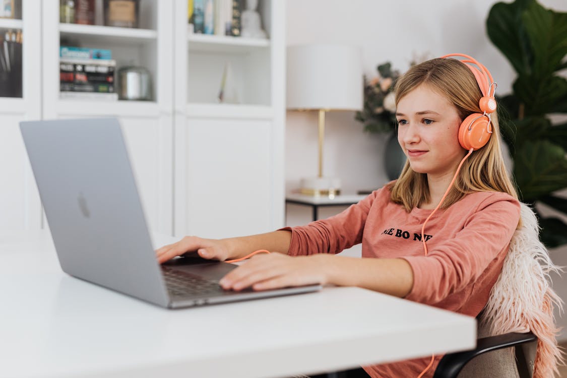 Free Young girl focused on using laptop while wearing headphones indoors. Stock Photo